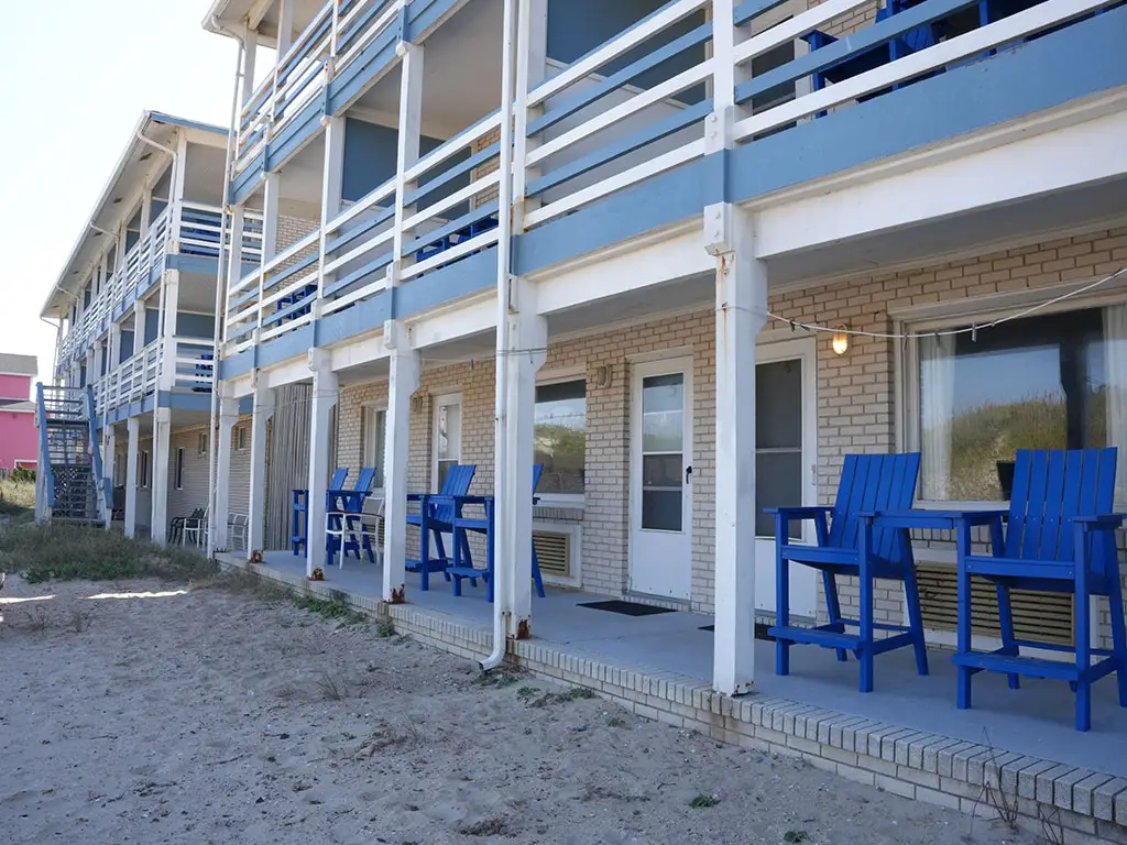 A two-storey building with blue balconies and white railings, with blue chairs on the ground-floor patio facing a sandy area
