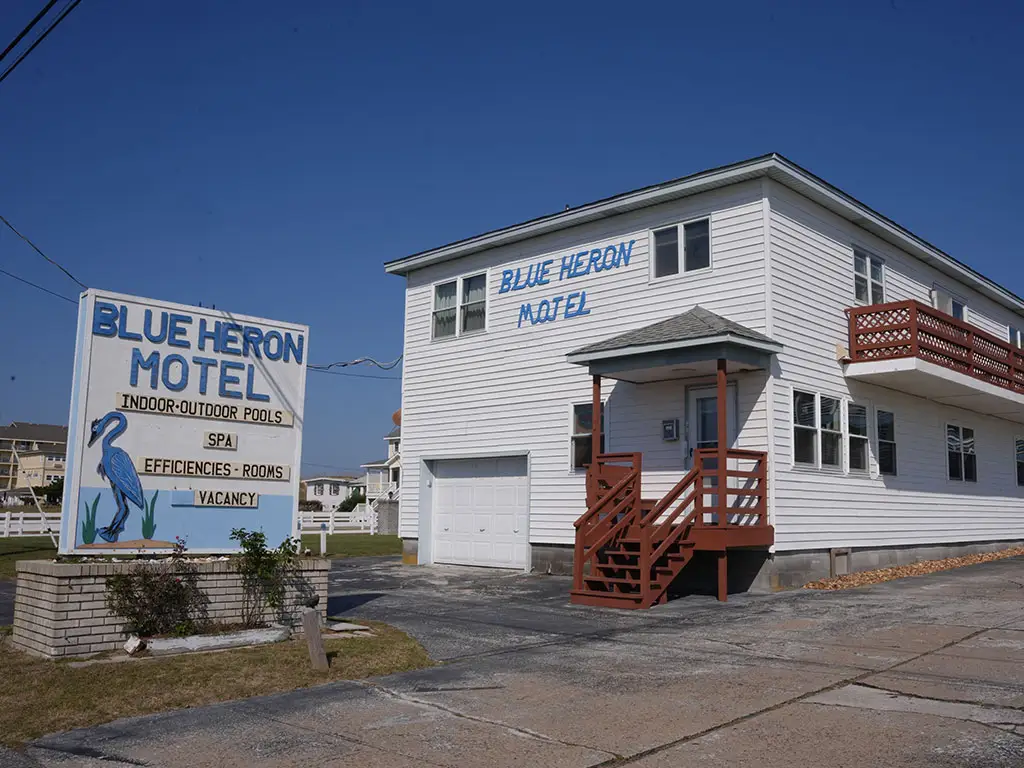 The white two-storey Blue Heron Motel building with a brown-railed balcony and a sign advertising indoor-outdoor pools, a spa, efficiencies, rooms and vacancy