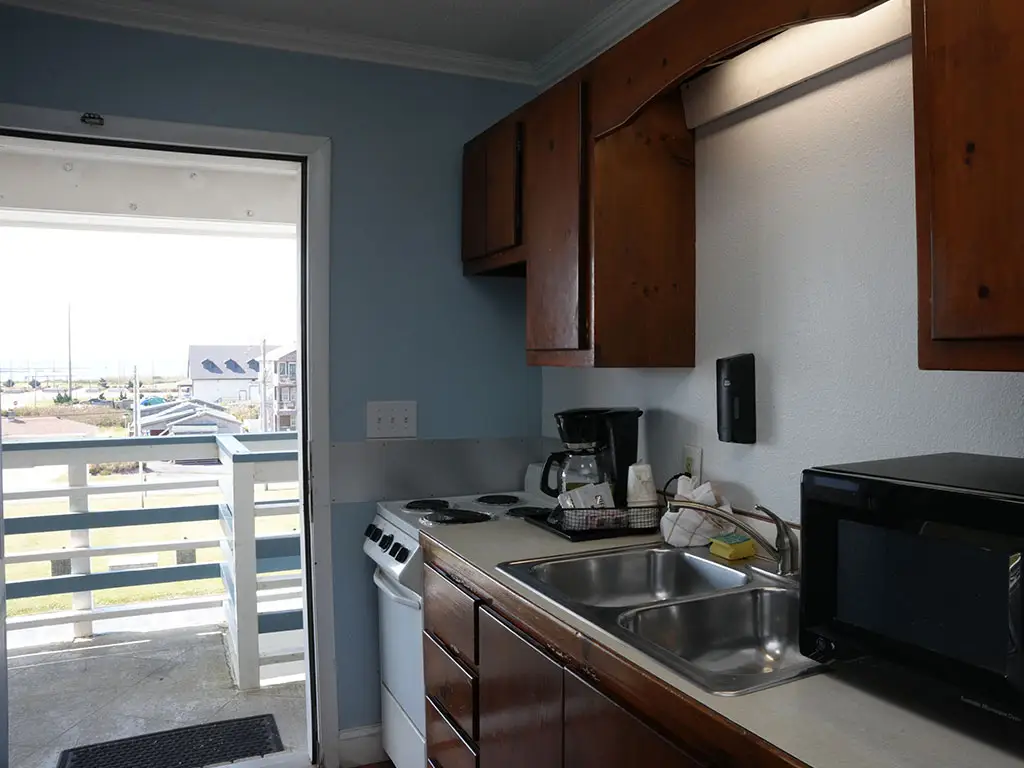 A kitchenette with a double sink, a microwave, a coffee maker and a stove, with an open door leading to a balcony overlooking buildings and the ocean