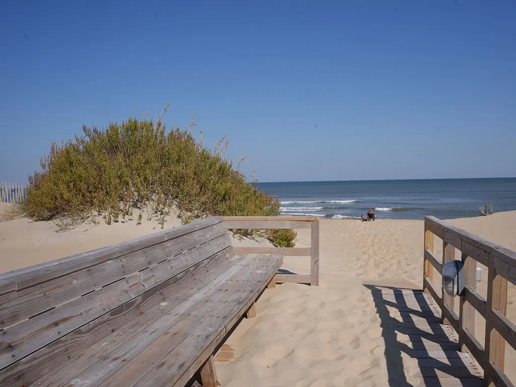A wooden bench and boardwalk lead to a sandy beach with a large green bush and two people near the ocean