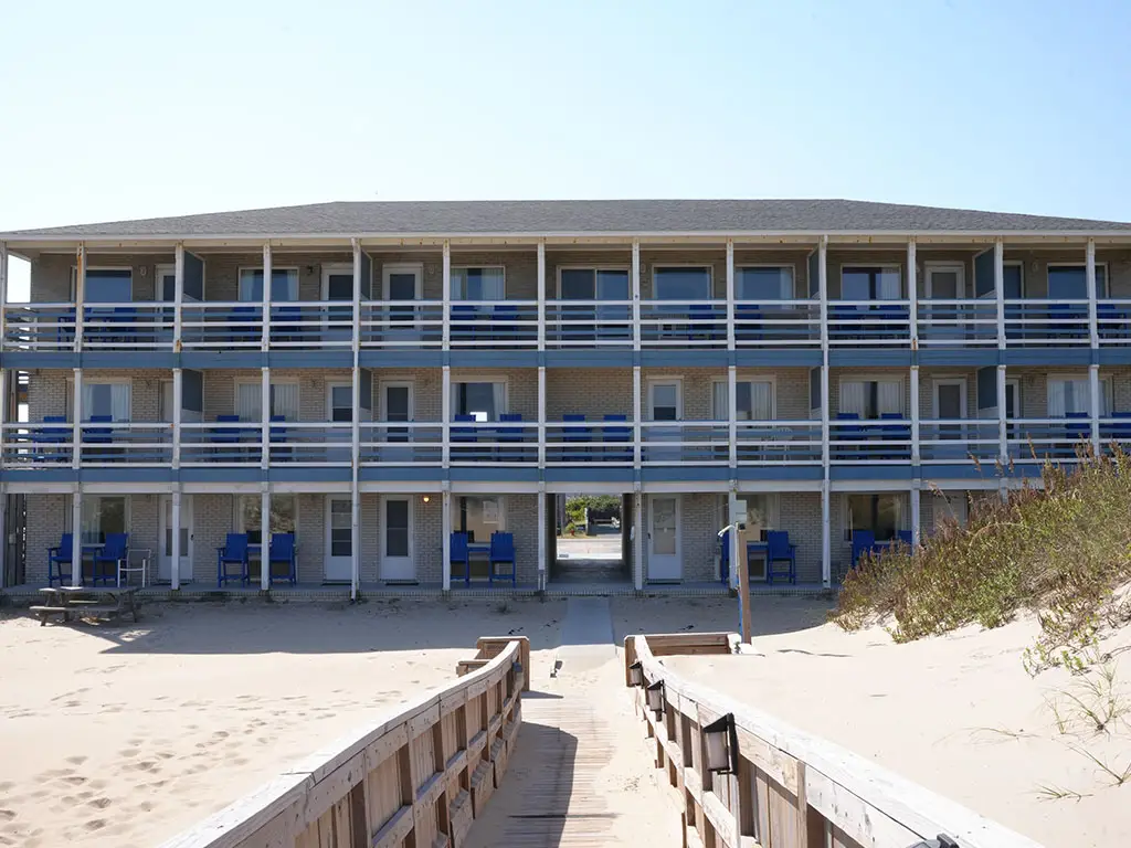 A three-storey hotel building with blue railings and white balconies, seen from a wooden boardwalk on a sandy beach