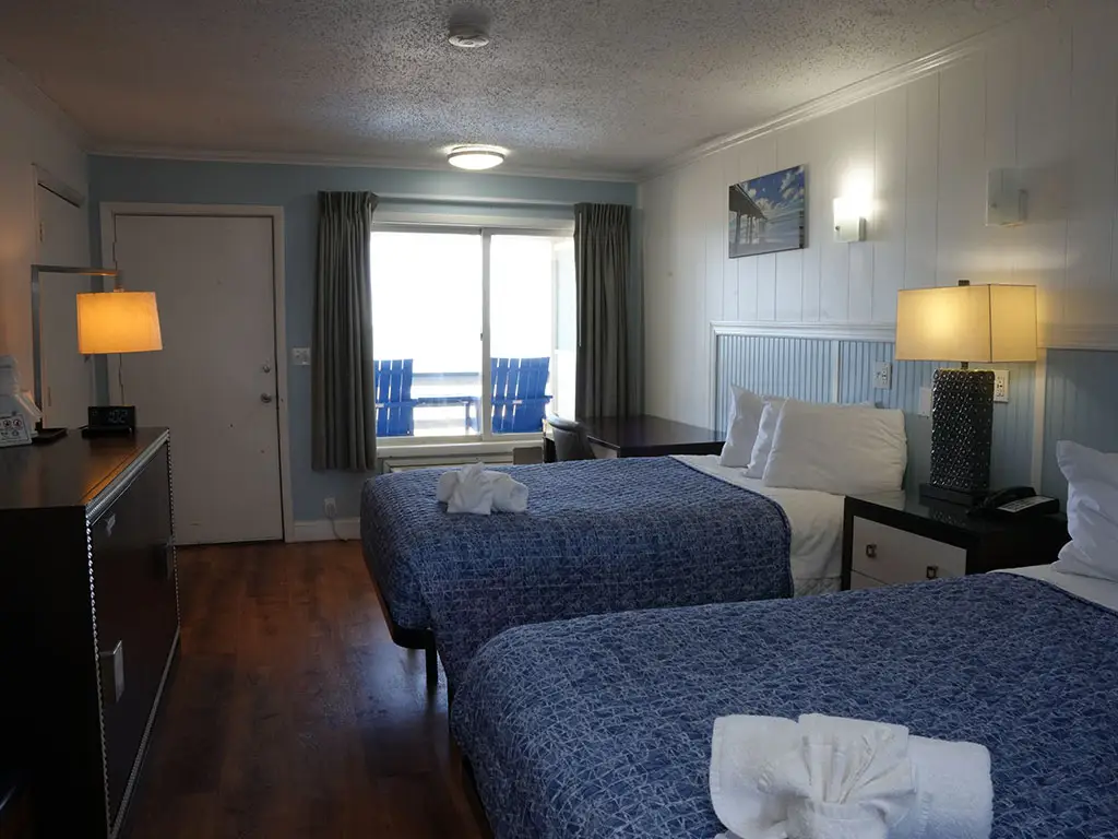 A guest room with two beds with blue patterned bedspreads, a dark wood dresser, and a sliding glass door leading to a balcony with two blue chairs