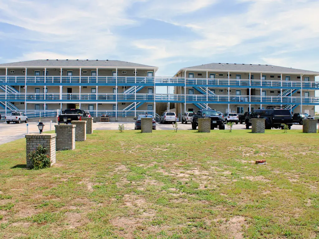 Two grey two-storey buildings with blue and white exterior walkways are separated by a connecting walkway, with a car park and grassy area in front