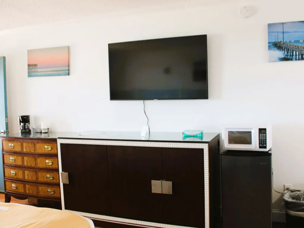 A hotel room with a wall-mounted TV, a dark wood dresser, a dark wood cabinet, a microwave and a mini-fridge