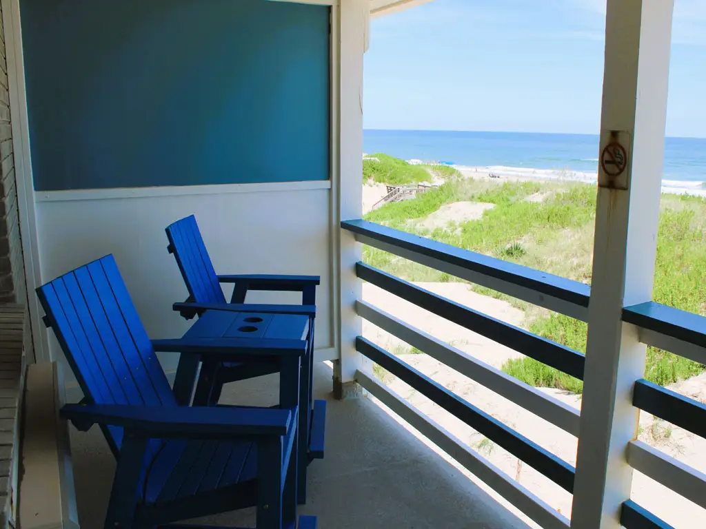A balcony with two blue Adirondack chairs and a small table, overlooking a sandy beach and the ocean