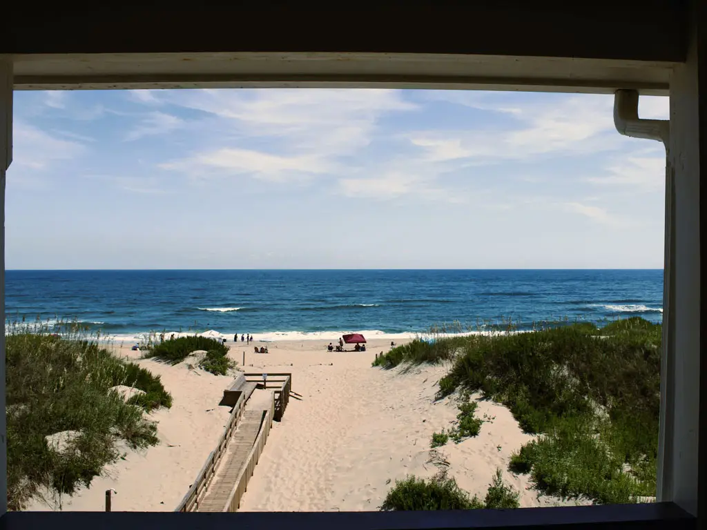 A view of a sandy beach with scattered people, green dunes and the ocean under a partly cloudy sky, framed by a dark overhead beam and side posts