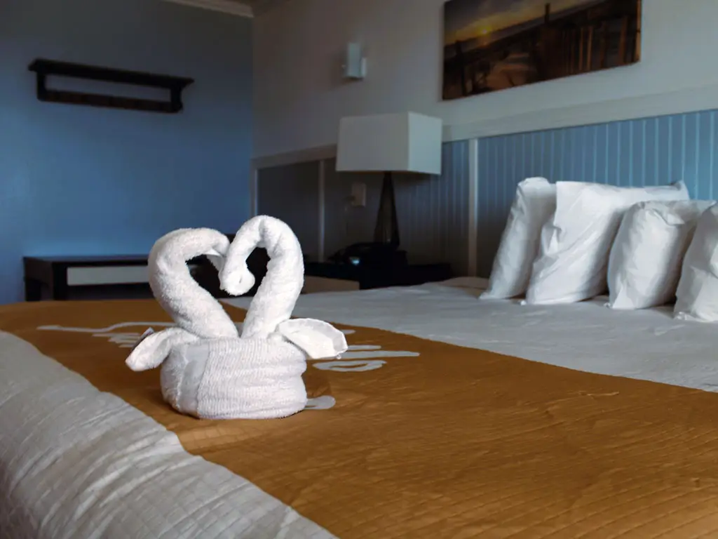 A white towel shaped like two swans forming a heart sits on a bed with a brown runner and white pillows