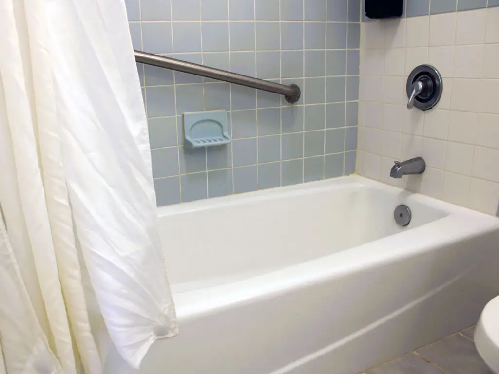 A white bathtub with a shower curtain, a grey grab bar and light blue tiled walls