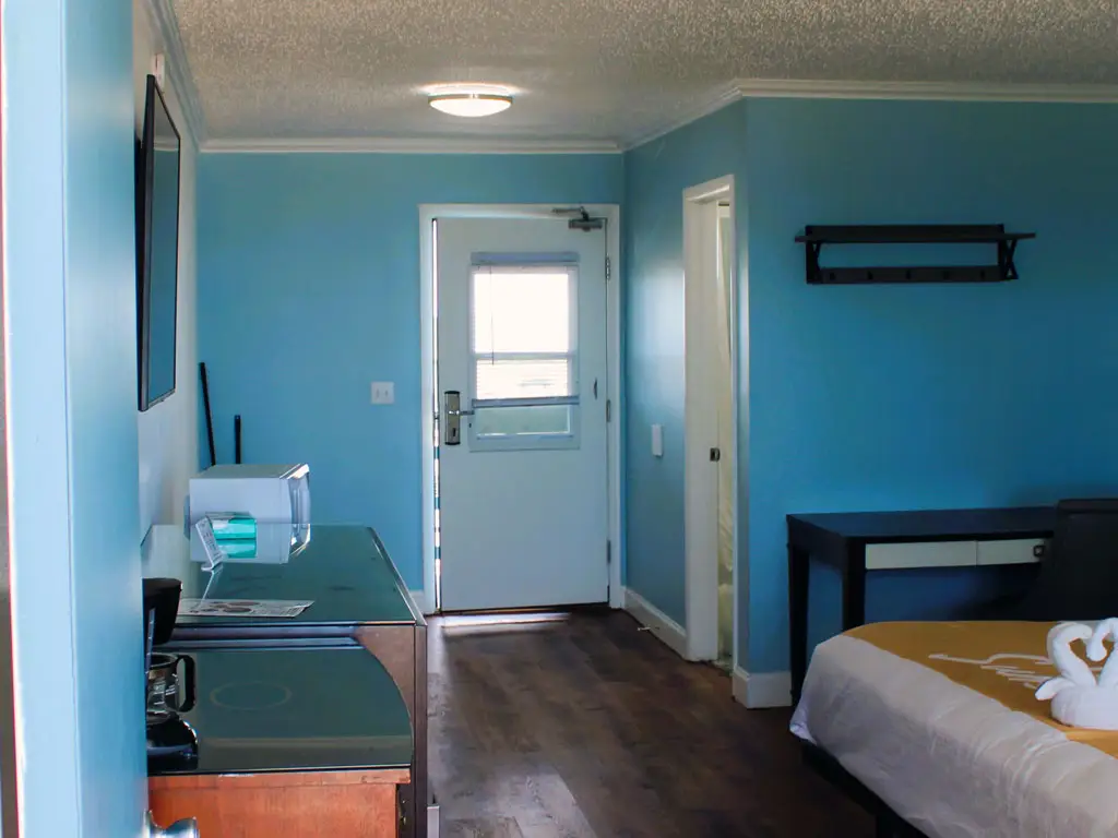 A hotel room with blue walls, a dark wood floor, a white door, a microwave, a coffee maker and a bed