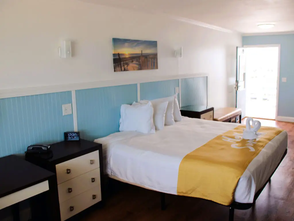 A guest room with a bed dressed in white bedding and a yellow runner, a bedside table, and an open door to the outside