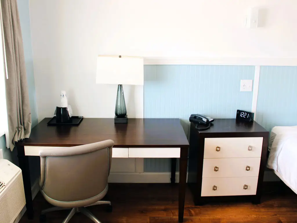 A dark wood desk with a grey office chair, a lamp, a tray with two bottles, and a bedside table with a phone and a digital clock