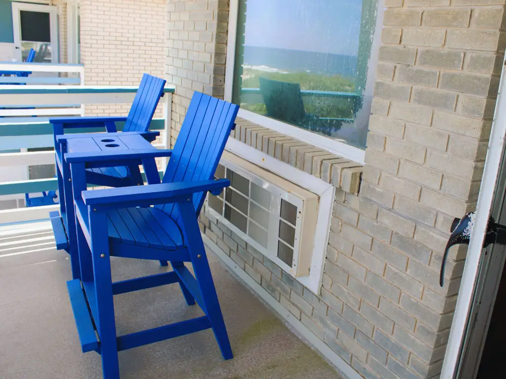 Two blue Adirondack-style chairs and a small table sit on a balcony overlooking the ocean, with a window air conditioner below the window