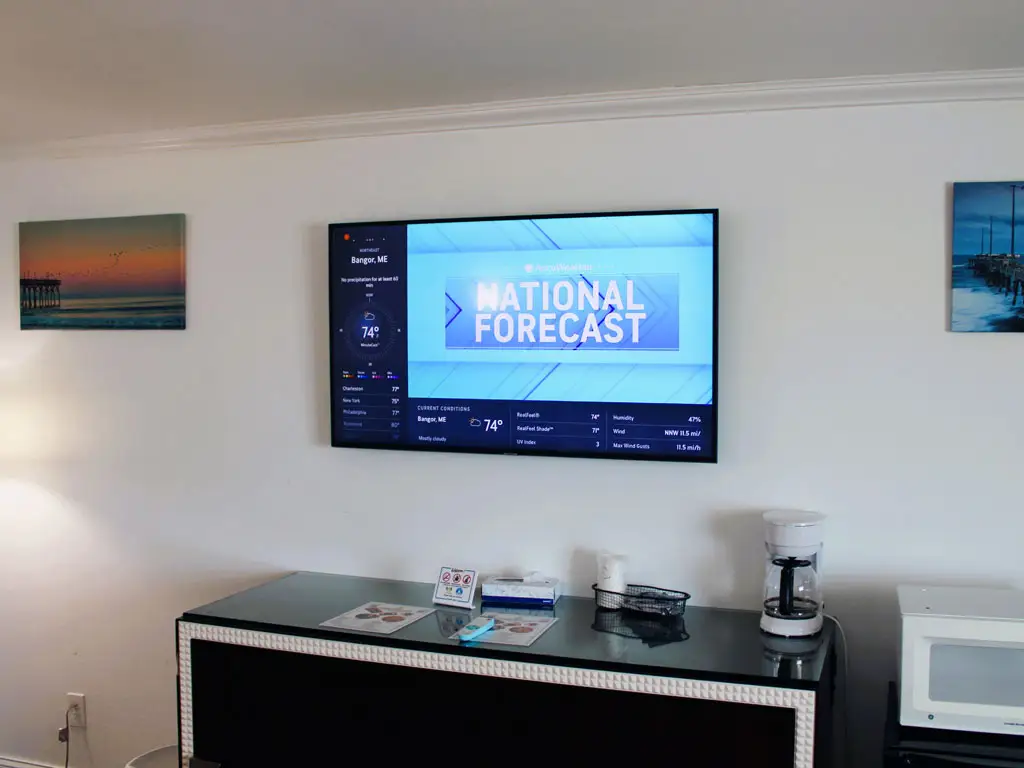 A wall-mounted TV displaying a weather forecast, with two framed pictures of a pier, a coffee maker and a microwave on a black cabinet