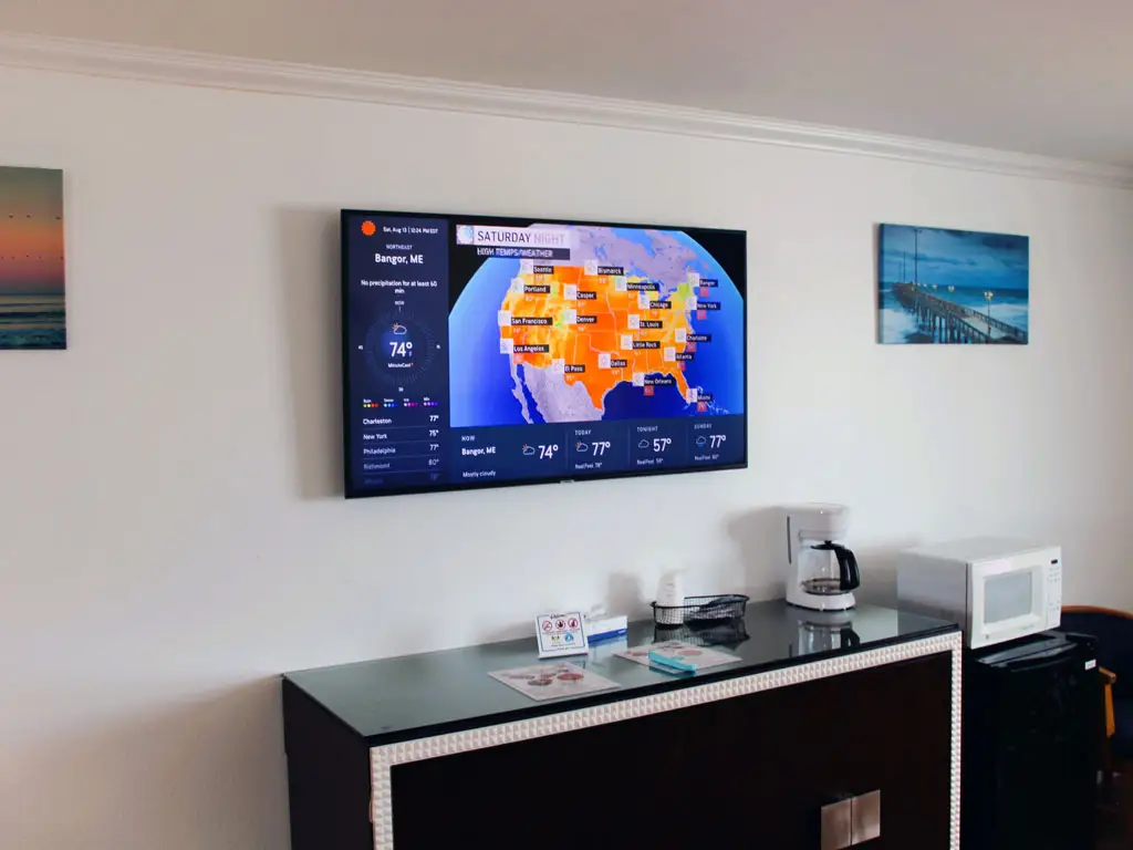 A wall-mounted TV displaying a weather map, a coffee maker, a microwave and a mini fridge on a dark cabinet