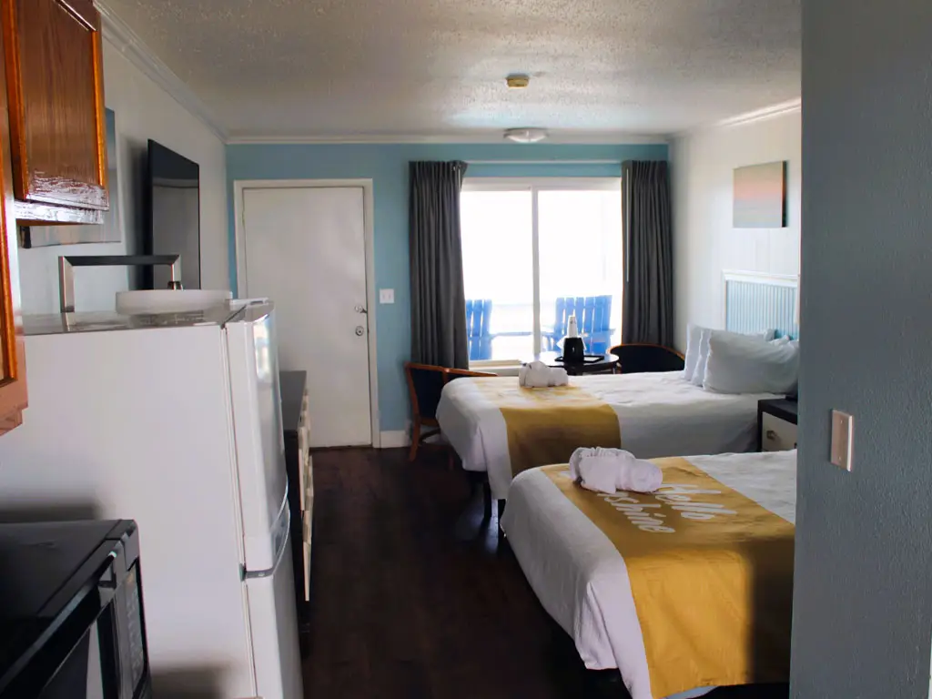 A hotel room with two beds, a small refrigerator, a microwave and a sliding glass door leading to a balcony with two blue chairs