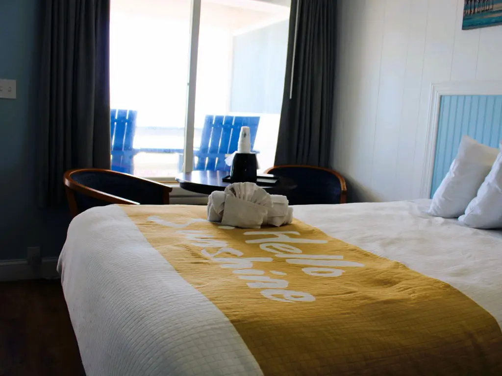 A hotel room with a bed in white and yellow bedding, two chairs and a table by a window overlooking a body of water
