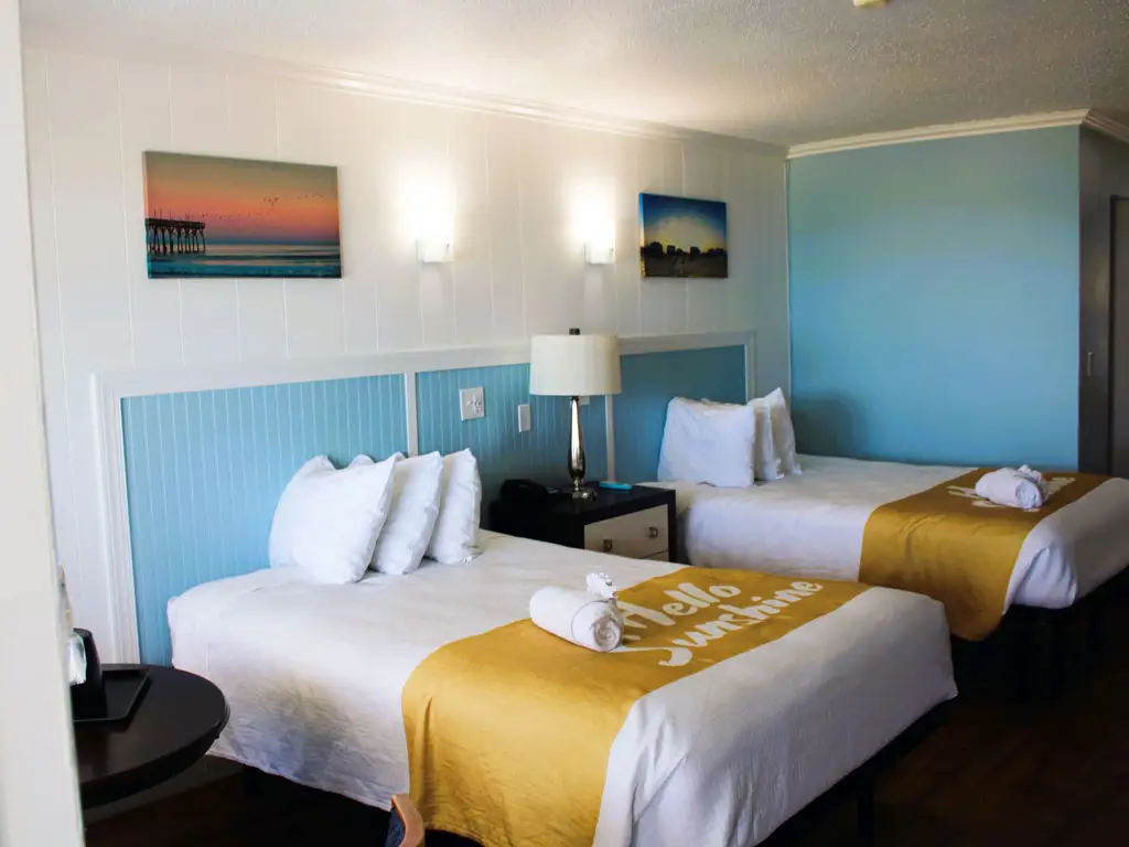 A guest room with two beds, each with a white duvet and a gold runner, and two wall-mounted pictures