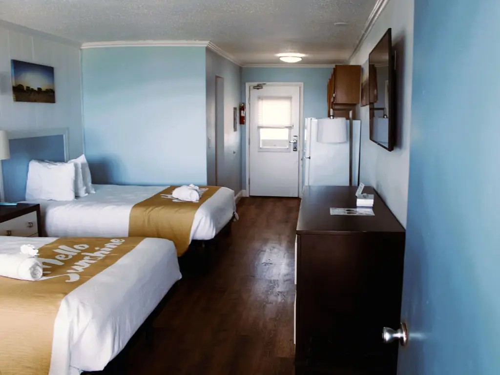 A guest room with two beds, each with white bedding and a gold runner, a wall-mounted television and a small refrigerator