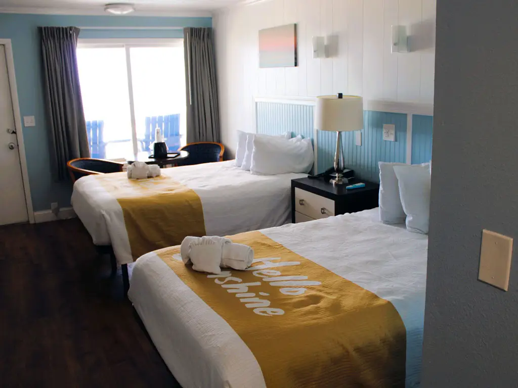 A hotel room with two beds, a bedside table with a lamp, and a sliding glass door leading to a balcony with two chairs