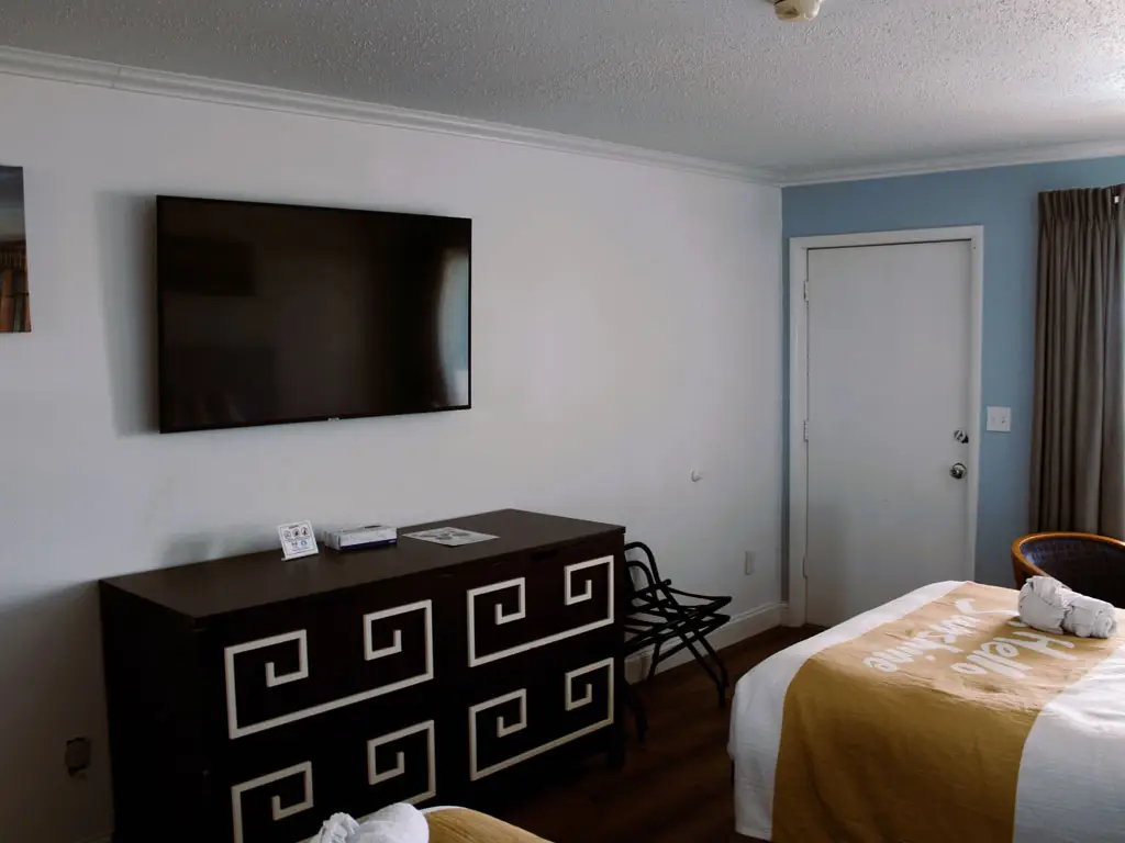 A guest room with a wall-mounted TV above a dark dresser, a white door and a bed with a yellow throw
