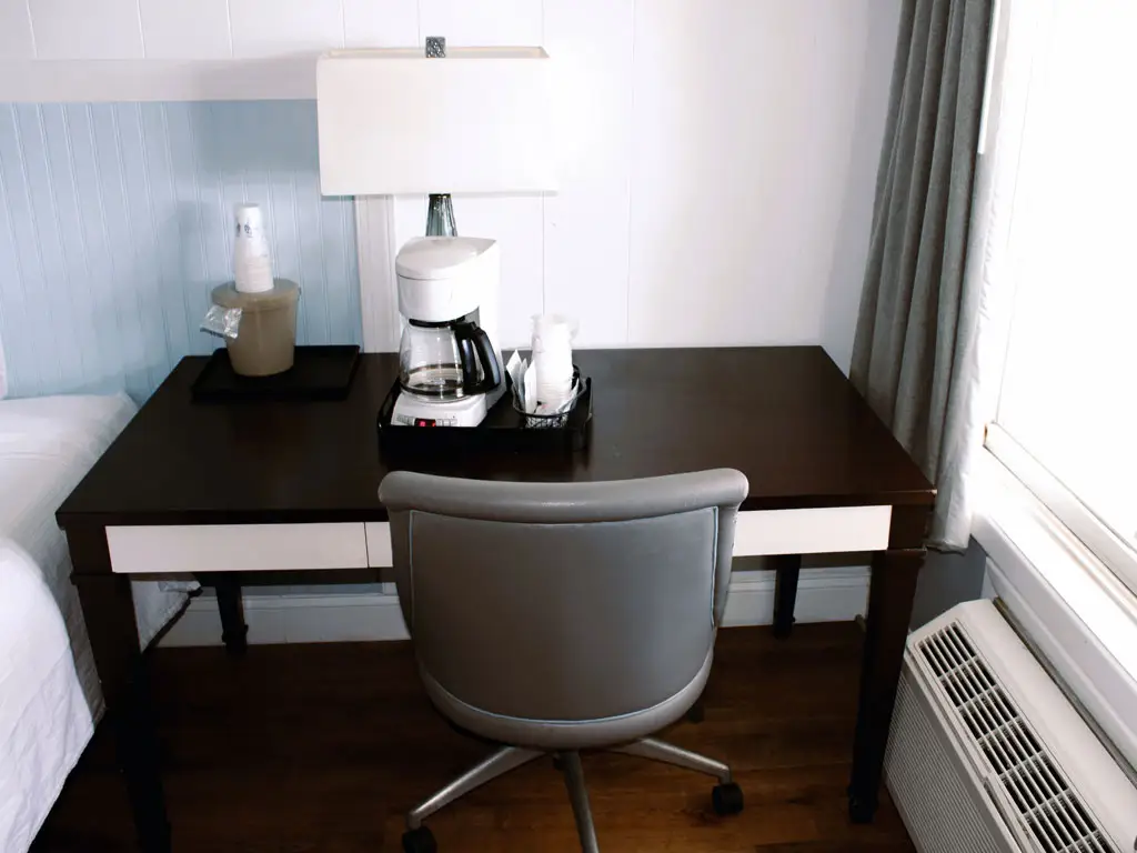 A dark wood desk with a coffee maker, a lamp, and a grey rolling chair, next to a bed and a window with an air conditioner