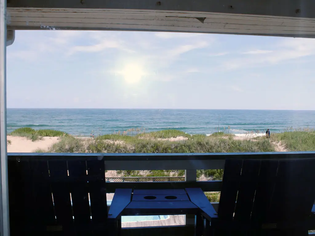 A view of the ocean and beach from a balcony with two dark Adirondack chairs and a small table