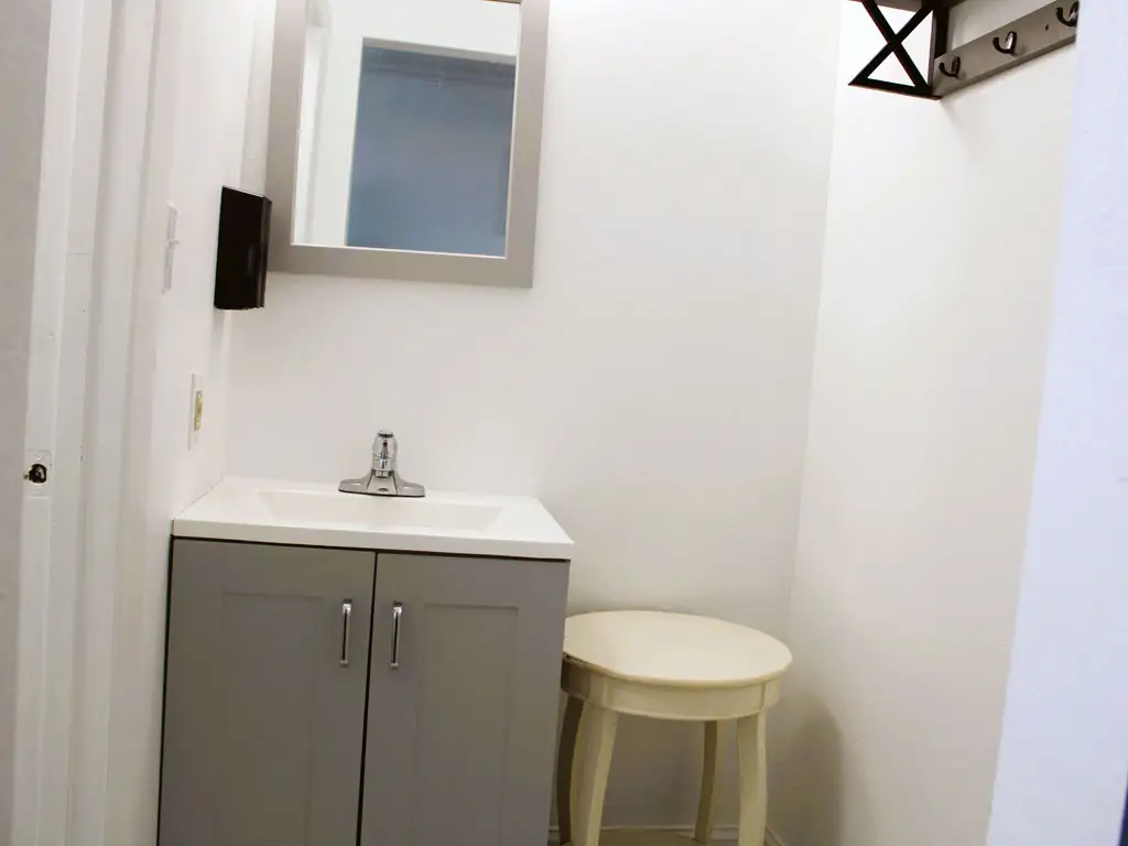 A small bathroom with a grey vanity, a white sink, a mirror, a black dispenser and a small round table
