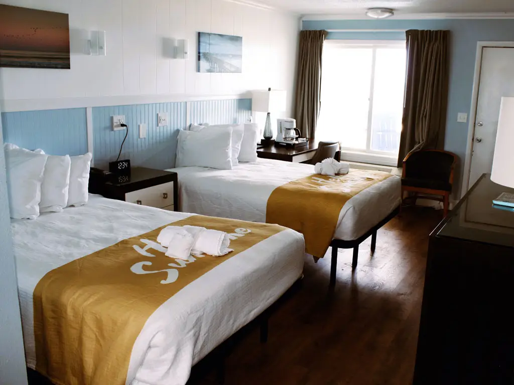 A hotel room with two beds, each with white bedding and a gold bed runner, and a window with brown curtains