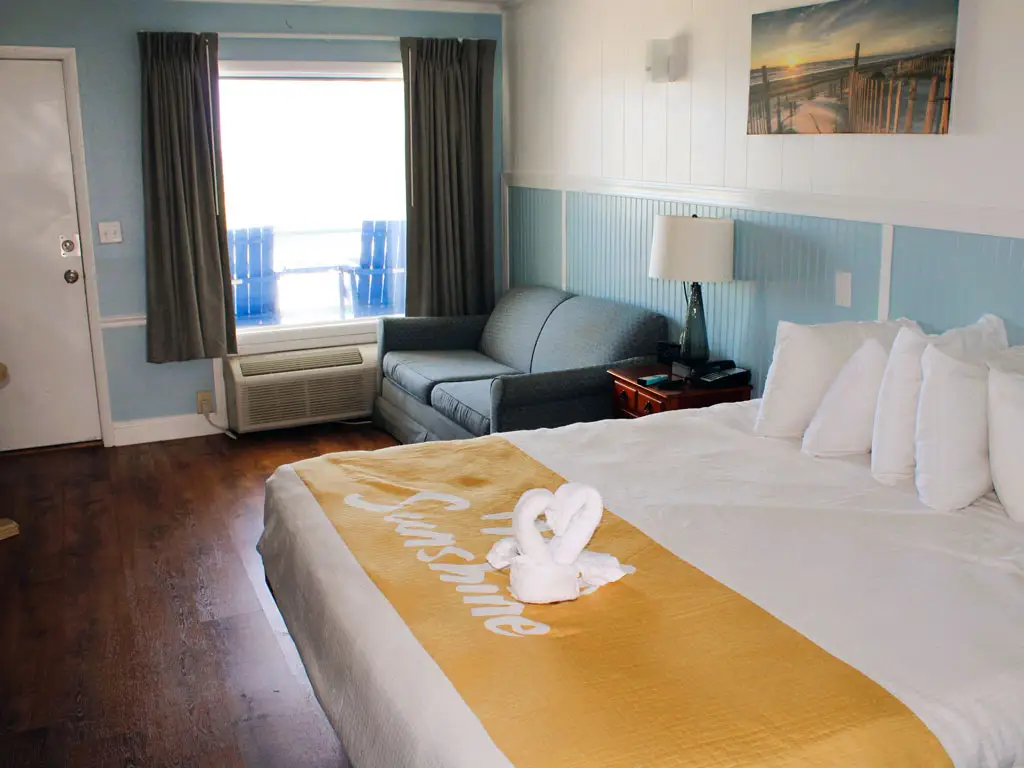 A hotel room with a bed, a sofa, a window with two blue chairs visible outside and a painting of a beach scene
