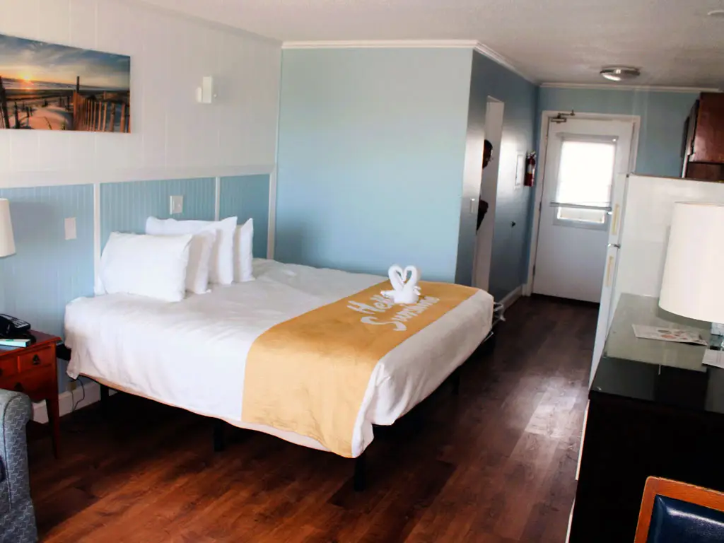 A guest room with a bed, a bedside table, a desk with a lamp, a refrigerator, and a door leading outside
