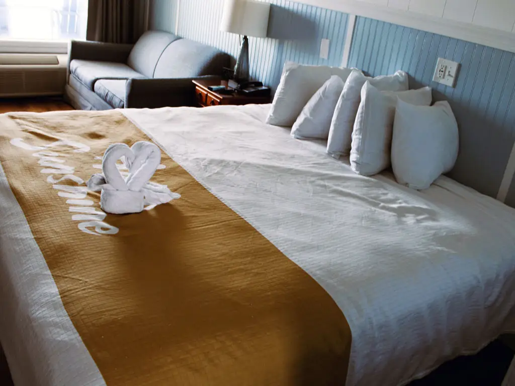 A bed with white bedding and a gold runner with "Sunshine" written on it, and a towel folded into a heart shape, with a sofa in the background