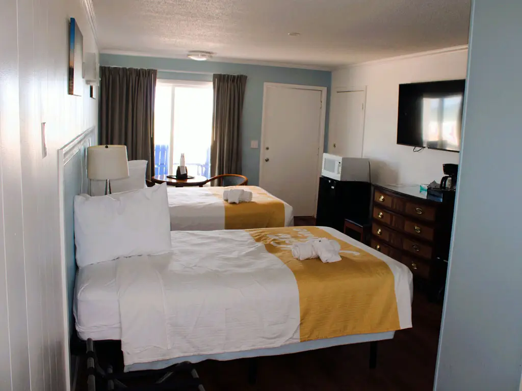 A guest room with two beds, a small table with two chairs, a microwave, a coffee maker and a wall-mounted television