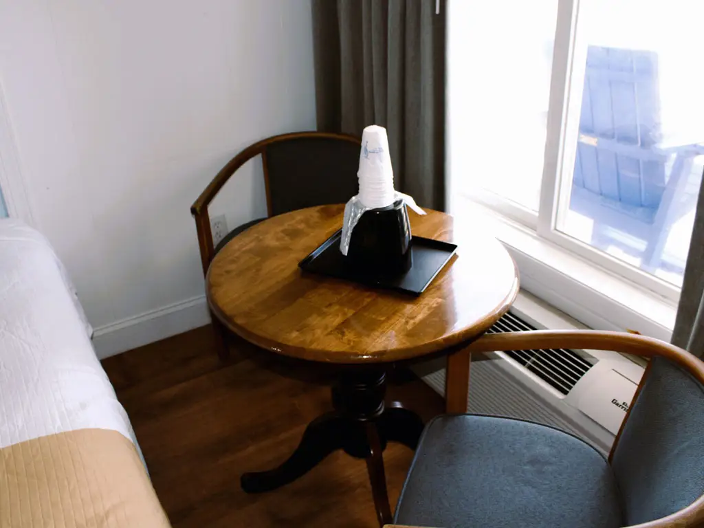 A round wooden table with two chairs, a stack of paper cups and a black ice bucket, next to a window