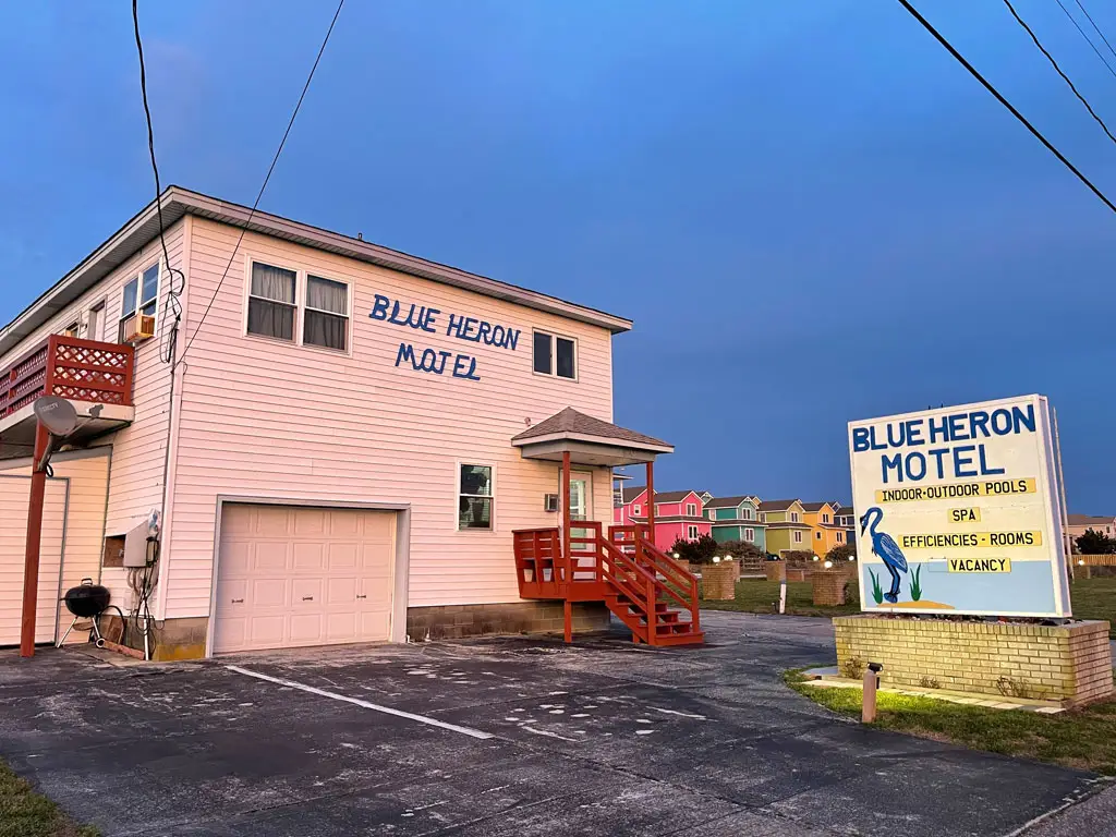 A white two-storey motel building with a red balcony and stairs, and a sign for "BLUE HERON MOTEL" in front of colourful houses