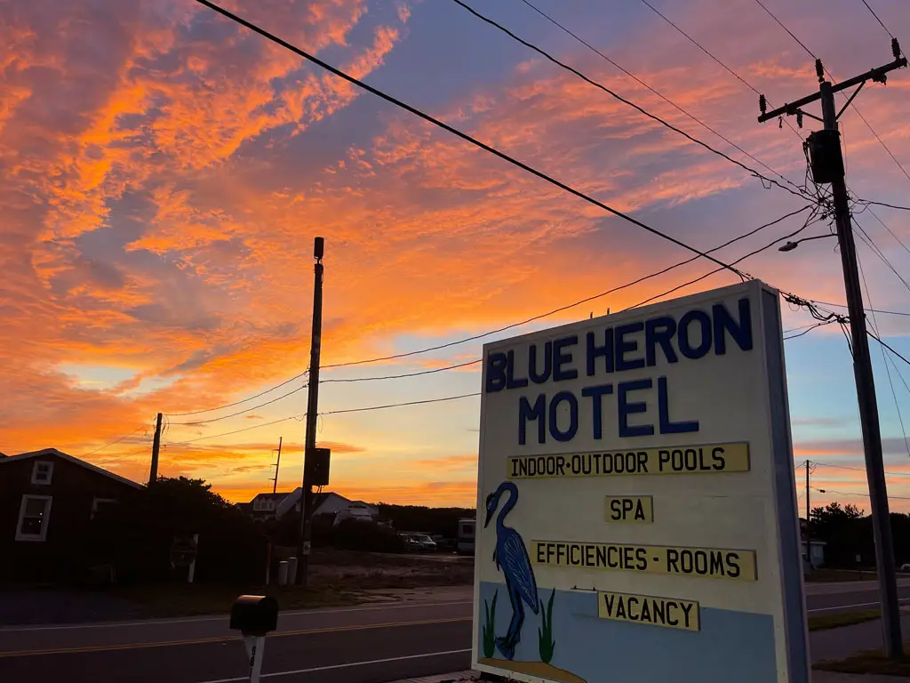 A motel sign for "BLUE HERON MOTEL" with "INDOOR-OUTDOOR POOLS", "SPA", "EFFICIENCIES - ROOMS" and "VACANCY" is visible against a vibrant orange and blue sunset sky
