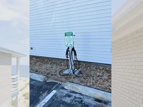 An electric vehicle charging station with two charging cables stands on a gravel patch next to a white-sided building