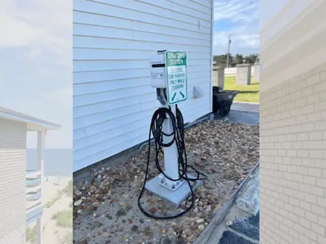 An electric vehicle charging station with a sign reading "Charging Station Electric Car Parking Only While Charging" is next to a white-sided building