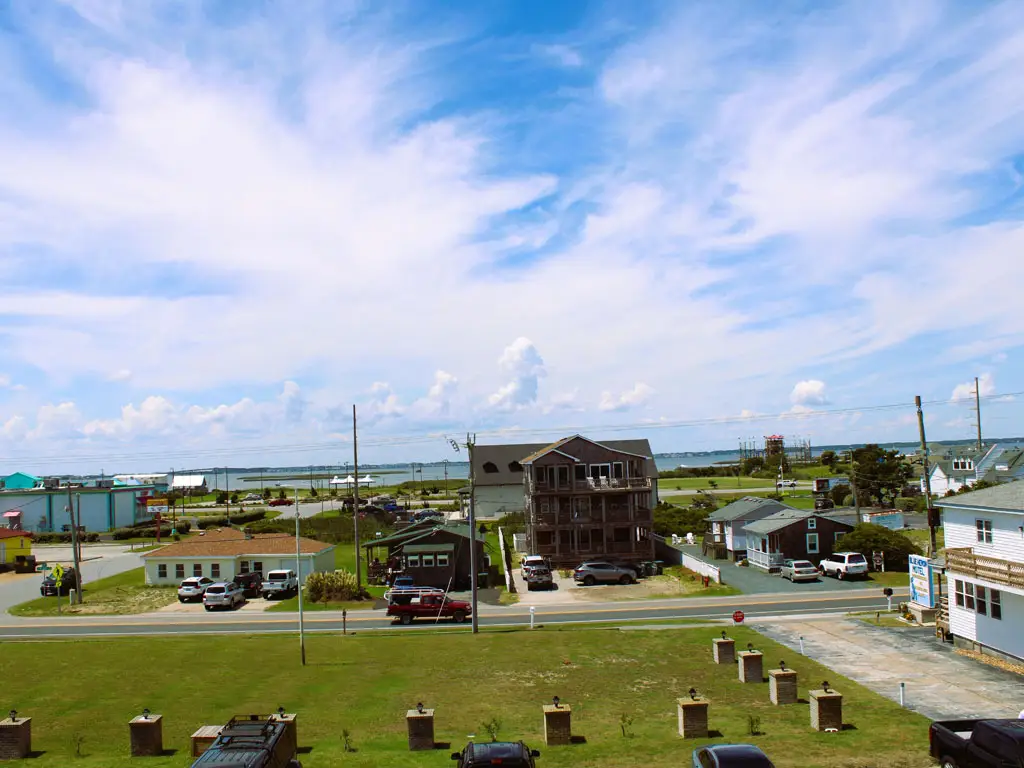 A coastal town scene with several buildings, cars, and a body of water under a blue sky with white clouds