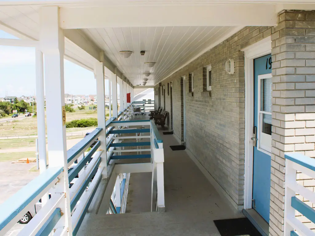 A long, covered outdoor walkway with a white railing and blue accents runs along a grey brick building with light blue doors