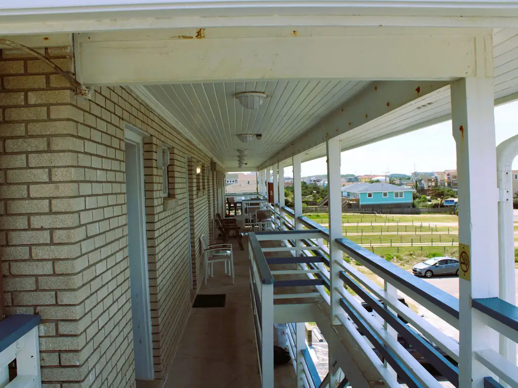A long, covered outdoor walkway with a brick wall on the left and a white railing on the right, overlooking a grassy field and distant houses