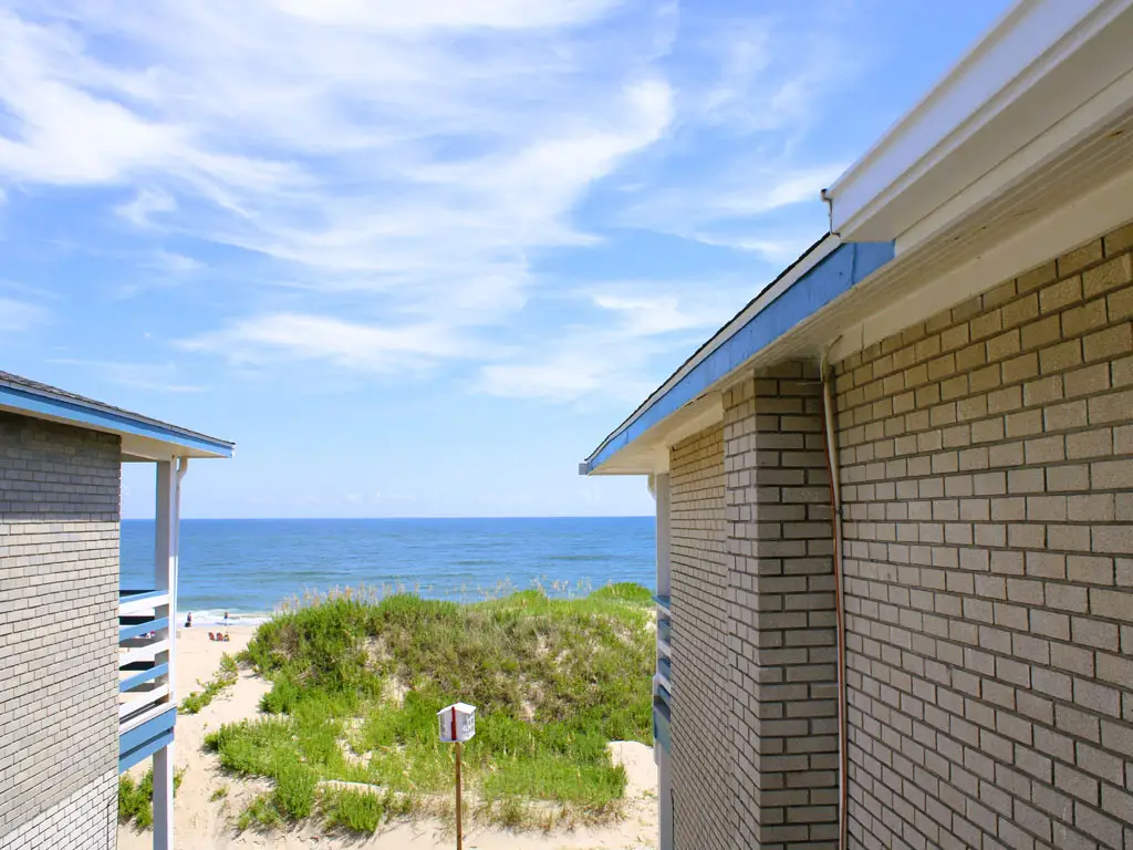 Two brick buildings frame a view of a sandy beach, dunes and the ocean under a blue sky with wispy clouds