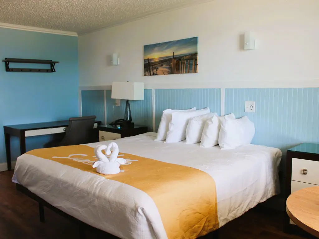 A hotel room with a bed featuring a gold runner and two towel swans, a desk and chair, and light blue wainscoting