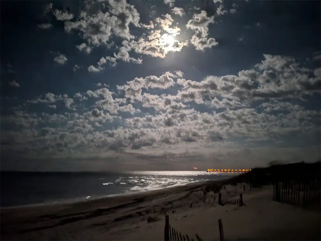 A moonlit beach scene with waves reflecting light, sand dunes, and a distant illuminated pier under a cloudy sky
