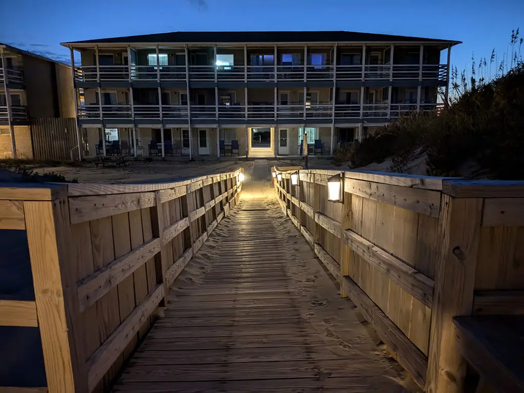A wooden boardwalk with lights on the railing leads to a three-storey building with balconies, seen at dusk