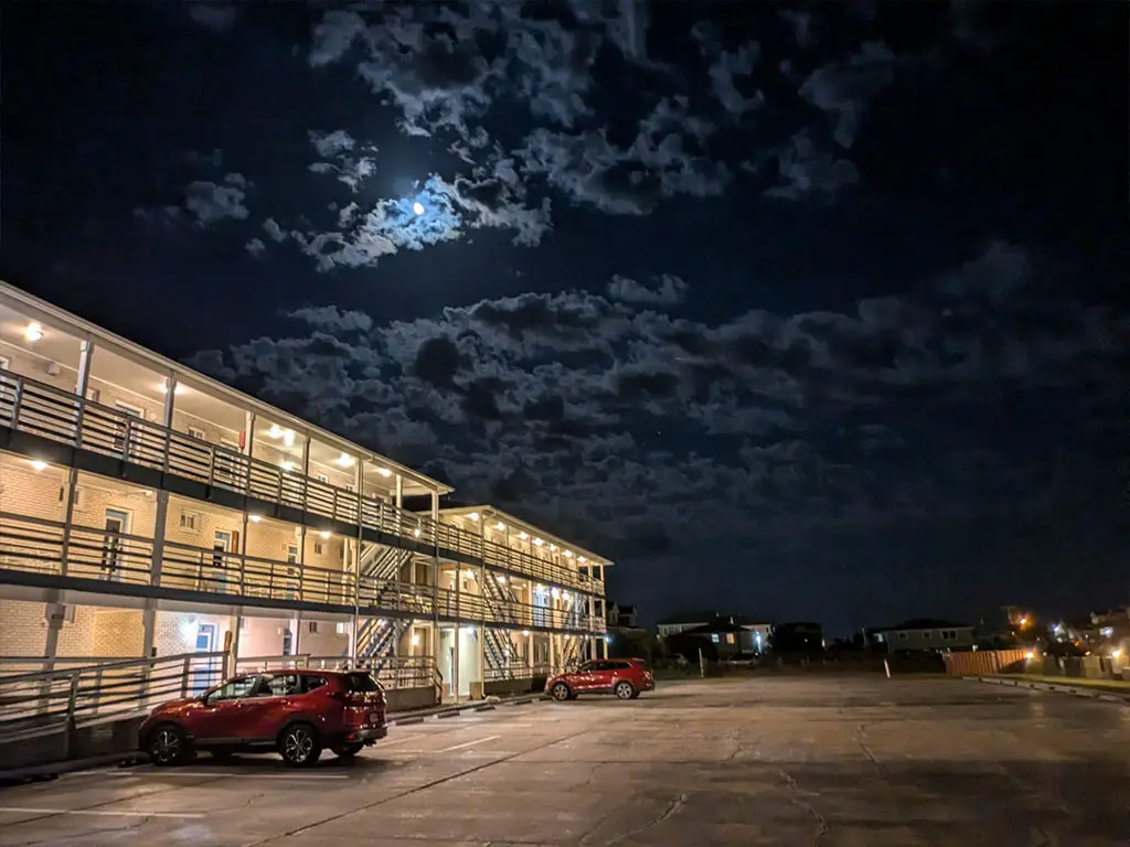 A two-storey motel building with external walkways and railings, a parking lot with two red cars, and a cloudy night sky with a visible moon
